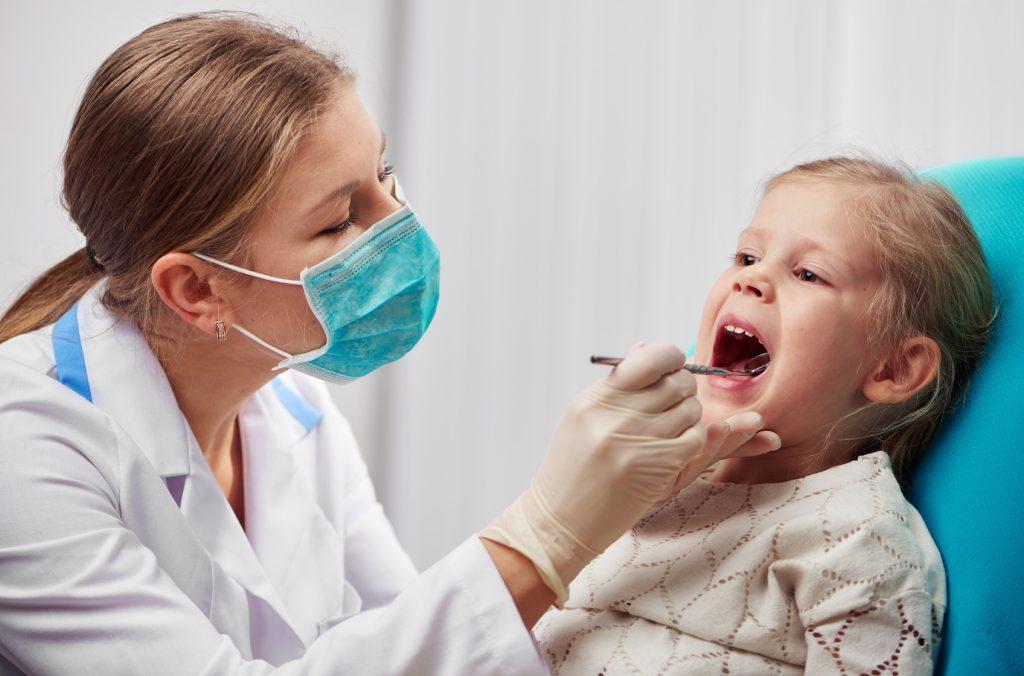 Woman dentist in mask doing teeth checkup of little girl in dental room. Health care and medicine concept.
