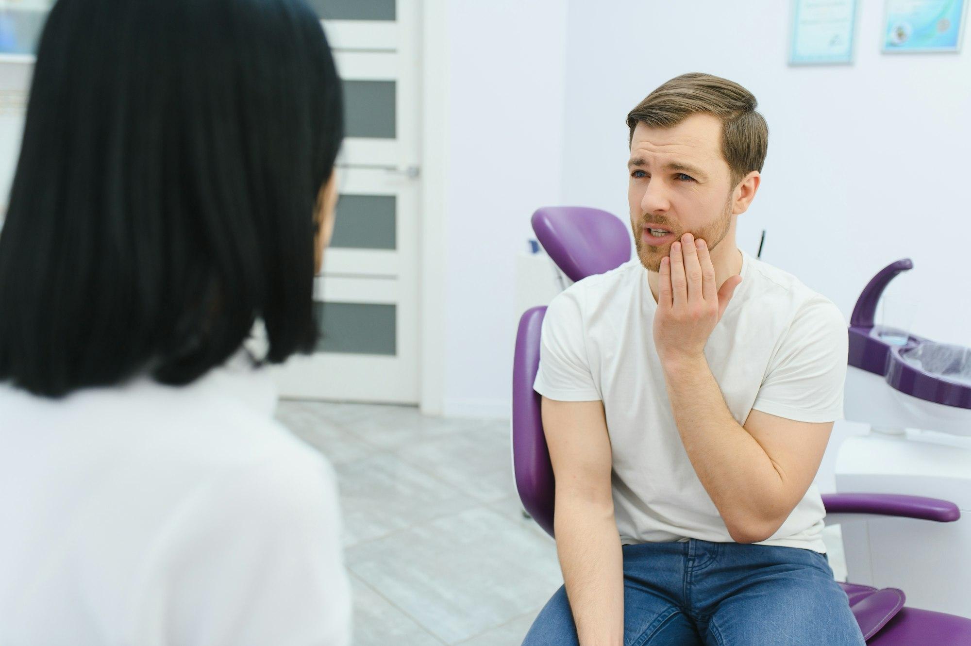 Sad man sitting in dental chair suffering from acute tooth pain, rubbing his cheek, close up.