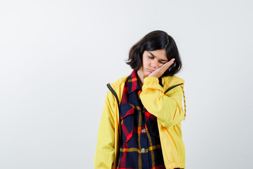 little girl pillowing face on her hand in checked shirt, jacket and looking sleepy front view