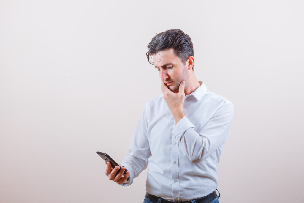 Young man in shirt, jeans looking at mobile phone and looking pensive with a toothache , front view.