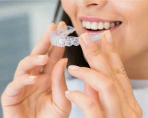 A close-up of a smiling woman holding a clear orthodontic aligner (such as SureSmile or Invisalign) near her mouth. Her clean, natural nails and white, straight teeth are visible, and she appears to be about to place the aligner onto her upper teeth. The background is softly blurred, keeping the focus on the aligner and her smile.