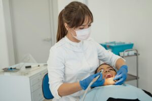 A female dentist wearing a white coat, face mask, and blue gloves performs a dental procedure on a female patient lying in a dental chair. The patient’s mouth is open while the dentist uses dental tools under a focused examination light. The clinical setting includes dental equipment and storage carts in the background.